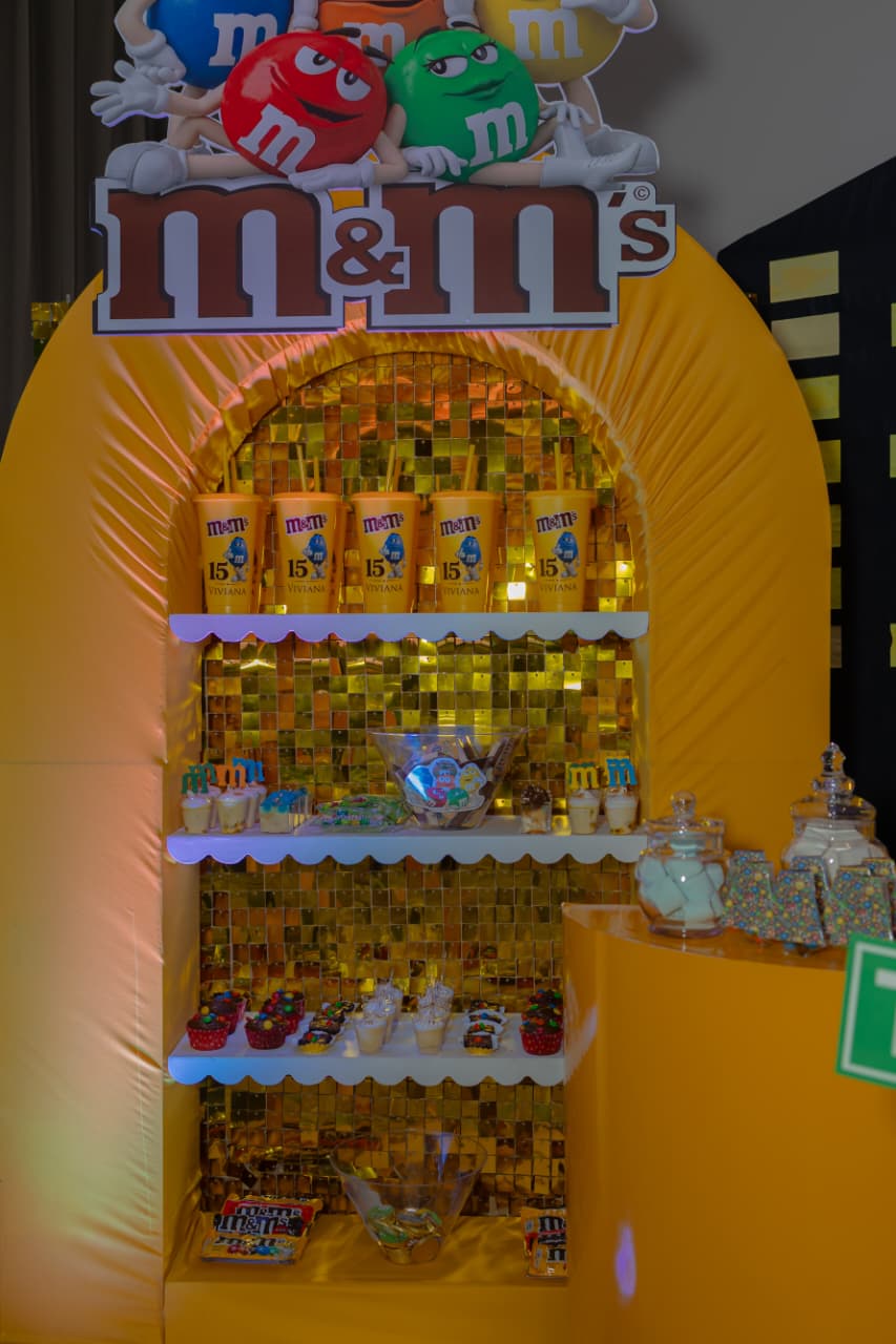 Estantería dorada de M&M’s con vasos personalizados, cupcakes y dulces