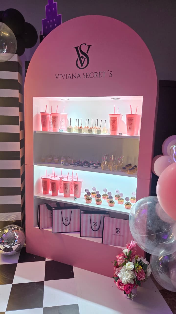 Barra de postres con arco rosado estilo Victoria’s Secret, vasos personalizados y cupcakes