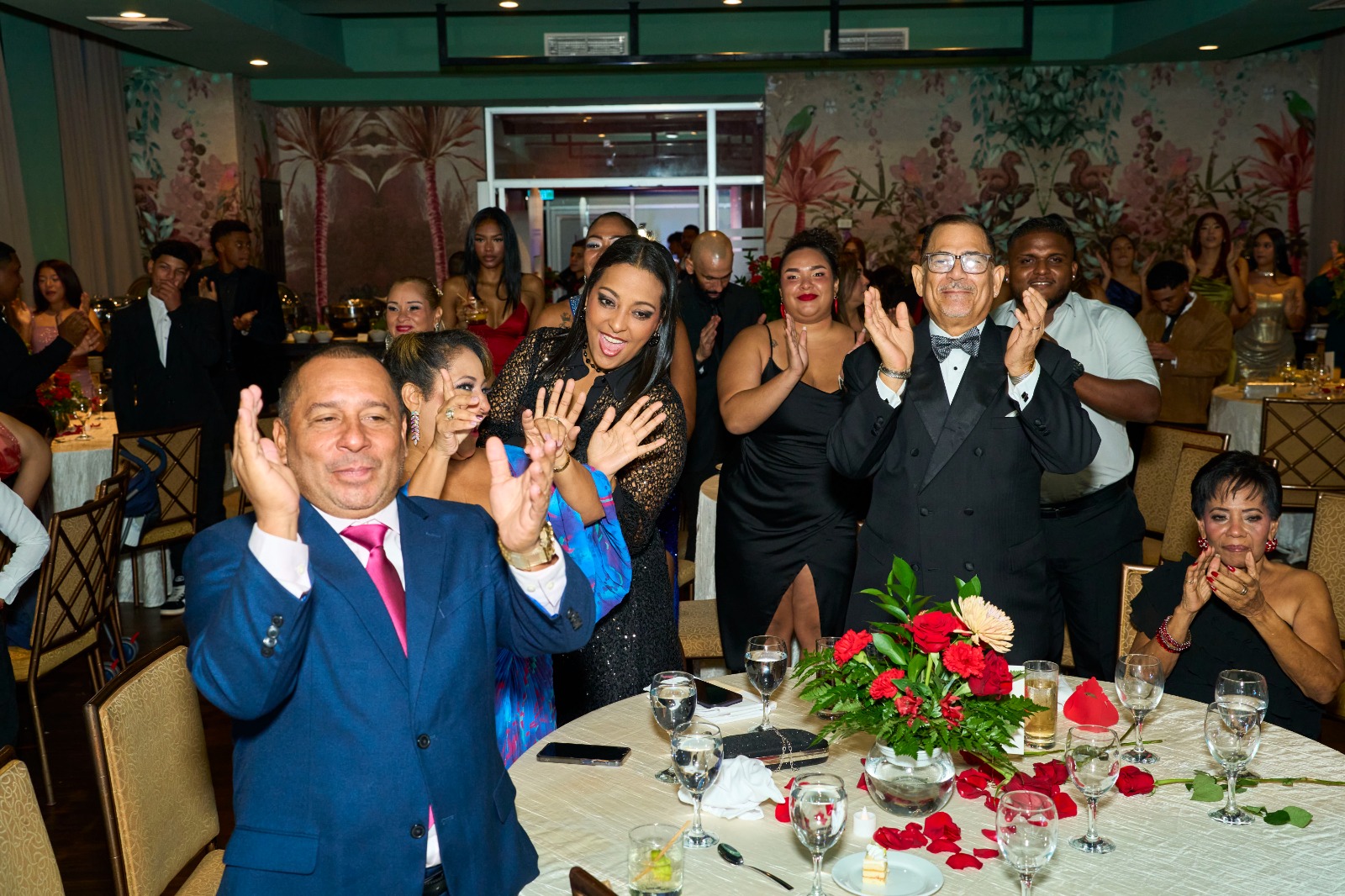 Invitados de gala aplaudiendo de pie en una celebración producida por Gloobeland, con mesas vestidas y centros de rosas rojas